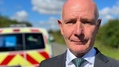 Darryl Preston, standing outside in front of a marked vehicle. In the background is a grassy verge and shrubbery next to a road. He is looking directly at the camera and is wearing a grey suit jacket, white shirt and green polka dot tie.