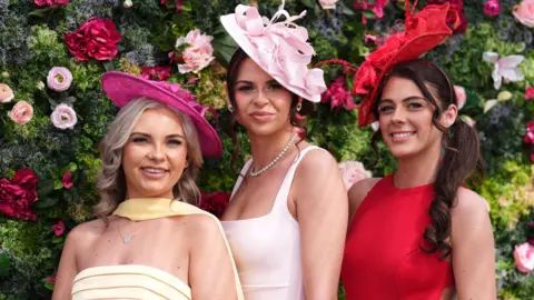 PA Media Three women stand in front of a wall of flowers. One wears a red dress and red fascinator hat, one wears a yellow dress and shocking pink hat and one wears a pastel pink fascinator and dress