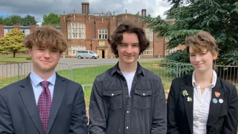 Henry Godfrey-Evans/BBC Three students stood side-by-side a few metres in front of a mass of bricks and a large school building looming in the background.