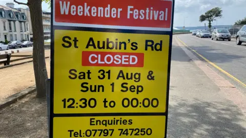 BBC A yellow sign with a red block at the top saying 'Weekender Festival' and details of the road closure. Behind is a walkway, a road on either side with queues of cars, on a grey cloudy day.