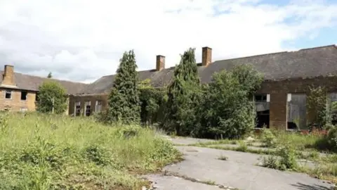 A photograph of the empty building that shows damaged windows and overgrown weeds outside