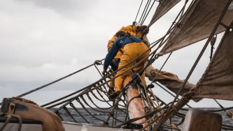 Kim and Del Hogg Crew members in yellow wet weather gear work the rigging on the bow of a traditional tall ship. 