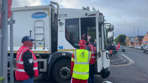 A large white bin lorry is stationary on a road. three people are standing next to it. The cab door is open. One man in a pink Unite the union jacket is talking to the driver. Behind him are two other men. One is in a pink unite the union jacket with a hat on and mask over his nose and mouth. The other has a yellow unite jacket on, with a red backwards cap