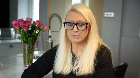 A blonde woman with dark-rimmed glasses, wearing black sits at a kitchen island with pink roses in the background. She looks into the camera with a serious expression on her face.