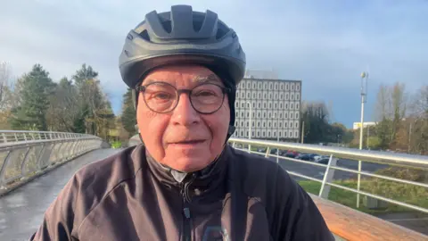 Paul Haughey is standing over his bike and wearing a black helmet. He's on a bridge and Marlborough Building is in the background.