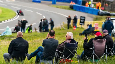 CLASSIC TT Spectators sitting in a row in camp chairs watch a motorcycle round a bend on the Mountain Road. They are on a bank of green grass and there are blue crash barriers at the side of the road in the distance.