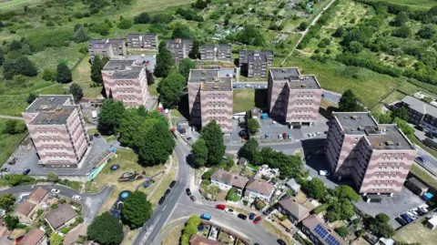 Aerials view of five seven-storey tower blocks of flats surrounded by various smaller buildings and green fields in the background