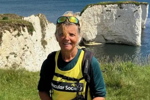 Scott Family A woman with blonde hair, and wearing reflective sunglasses on her head and a yellow Macular Society vest over a green top. She is standing on a coastal path, with the sea and a chalk promontory behind her.