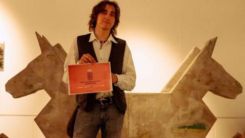 Artist Kamil Duda, in a brown waistcoat, white shirt and grey jeans, holds a red certificate in front of his artwork, a double-headed rocking horse made from a pale brown material