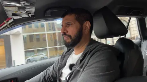 BBC/DAN BATER Akram Hussain Sharif, who has black hair and a beard and is wearing a grey hoodie, driving his taxi.