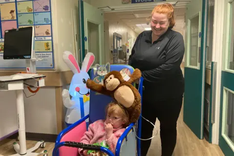A young woman with red hair pushes her young daughter in a wheelchair in a hospital corridor. The little girl has a tube attached to her face.