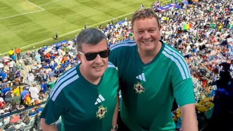 Two men wearing green Northern Ireland football shirts in a stadium, there are thousands of fans behind them.