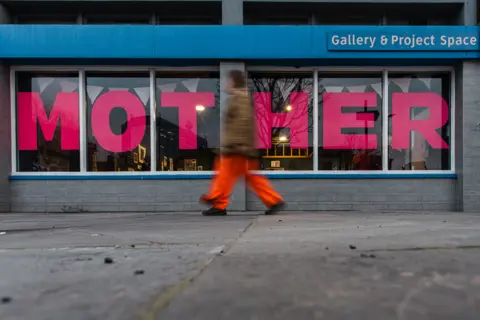 Neal Campbell blurred figure walking past a sign saying "mother" in the windows of an exhibition space