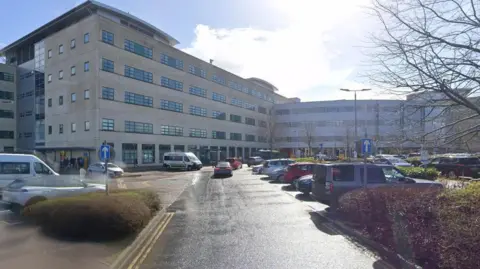 The front of the hospital on a sunny day - a large, modern building with lots of windows and a car park in front. 