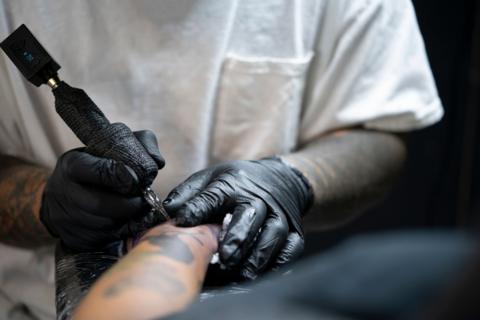 A tattoo artist wearing a white t-shirt and black latex gloves, holding a needle against a clients skin while tattooing