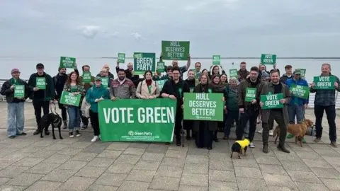 Members of the Hull and East Riding Green Party are gathered together standing in a crowd smiling at the camera. They're attending the party's campaign launch for Hull's local elections. Some of the members are holding up Green Party signs with their slogan which says 'Hull Deserves Better'. Some members have also brought their pet dogs to join the launch.