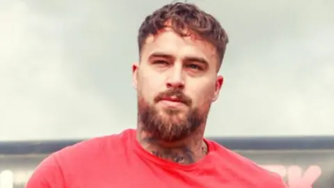 Jordan has short brown hair, which is cropped at the sides, a beard and a tattoo on his neck. He is wearing a red t-shirt.