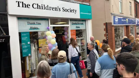 A charity shop with a large sign reading "The Children's Society." There are balloons beside the door and a crowd on nine people entering the shop. A woman and a man in mayoral chains stand beside the shop.