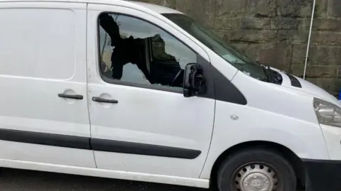 A white van on the street in daylight, with its window smashed.