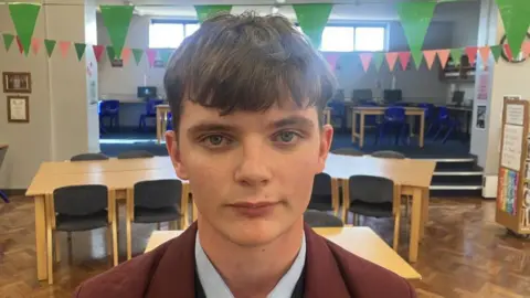 Caoimhin, a teenage boy with brown hair, a claret blazer and light blue shirt. There are brown tables and green bunting in the background.