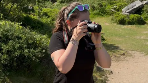 Jelly Boxall, with sunglasses on her head and her long, dark hair in a pony tail, is wearing a black t shirt and is taking a photo with her camera on Dartmoor. 