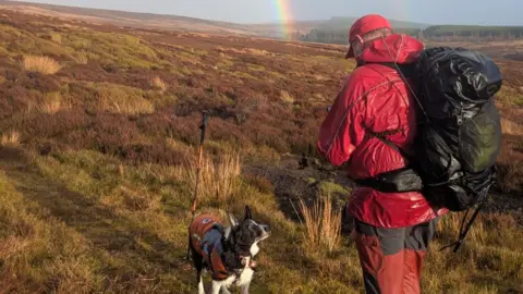 A black and white sheep dog looking up at a man dressed in red mountain rescue colours. They're standing on moorland and the bottom of a rainbow can be seen in the background.