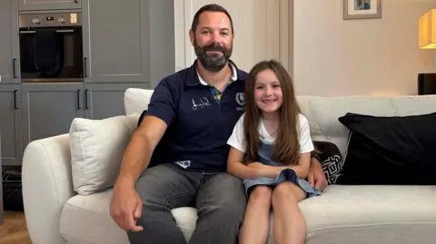 Perthshire Picture Agency/PA Paul Choat, 40, from Longniddry, is pictured with his daughter Skye after taking on the challenge of climbing all of Scotland’s tallest mountains. They are sitting on a beige couch in a domestic setting. Paul is wearing grey jeans and a blue polo shirt. His daughter is wearing a white t-shirt and a denim skirt. 