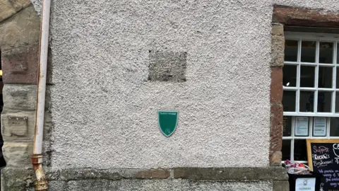 A faded white wall with a green sign. Above the sign is a blacked out area where a plaque once stood. A white sashed window sits to the right of the image, with stone work surrounding it. 