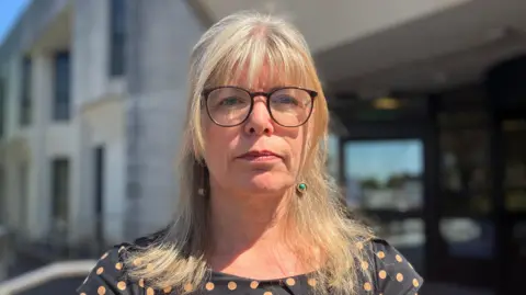 Liz looks directly at the camera. She has a long, straight blonde hair and a fringe and is wearing glasses and a black top with beige polka dots on it.