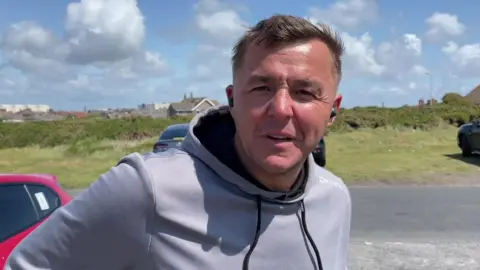 Antony Goldsmith is wearing a grey hoodie and wireless ear pods. He has short brown hair. He is standing in a car park in Barrow.