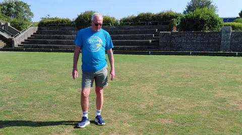 A man in a blue top with a fish round logo on it, green shorts, blue trainers with white socks is standing on a bowling green and looking down at the floor whilst frowning.