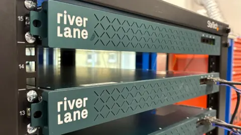 Ben Schofield/BBC A computer server rack. There are two identical-looking devices in the shot, which have "Riverlane" written on them. They are a deep green colour and have wires plugged into them.