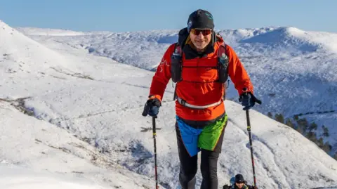 David Murch / Montane Spine Race Runner Chris Wright ascends up a snowy hill. He is wearing warm winter clothing with trail running poles and a backpack. It is a bright sunny day and he is wearing sunglasses.