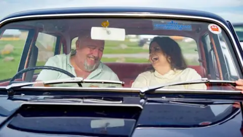ITV Cymru Wales / BBC Cymru Wales Jones and Speirs in an old navy vintage car laughing with each other. Jones, on the right, is in the passenger seat and is laughing whilst looking at Speirs. She wears a cream jumper and has brown hair. Speirs, is on the left, and wears a light green t-shirt and is looking down while laughing.