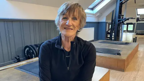 Leeanne Rinne/BBC Rachel Glenny looks straight into the camera wearing gym gear and sitting in a weightlifting room.