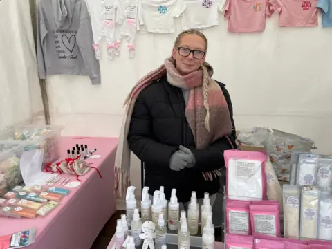 Samantha stands behind a table with bath products on. She is wrapped up warm in a black coat and gloves and scarf.