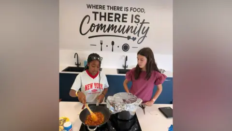 The Way Two girls stand over a hob as food is cooking. One girl is stirring food in a pan. Behind them on the wall is a painted message: Where there is food, there is community.