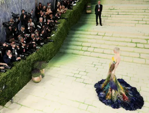 MG26/Getty Images for The Met Museum/Vogue Influencer Emma Chamberlain wears a multi-colored gown that resembles a painting. It begins with dark blue colors at the base before transforming into a yellow and teal abstraction. Her sleeves also have fringes down to the floor