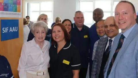 University Hospitals Coventry and Warwickshire A group of men and women standing in front of a unit and a wooden door. A blue sign on the door reads "SIF AAU". There is a patient, who is wearing a white blouse and beige trousers, and she has short white hair. The woman next to her is in dark blue nurse uniform. On the right are three men wearing suits.