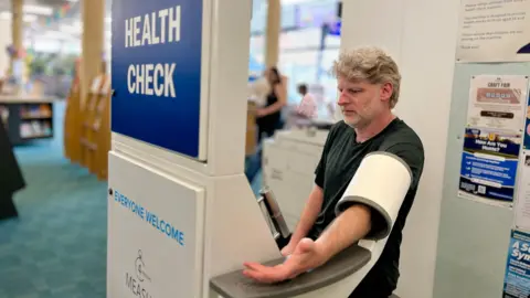 MARTIN GILES/BBC Michael Hadley has his arm through a blood pressure monitor cuff. It is attached to the machine and is a tube which has the monitor cuff inside it. It inflates as he rests his arm on a plastic ledge. The machine is labelled 'free health check', You can see some library shelves in the background