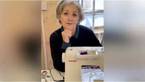 Contributed Lesley Partridge rests a hand on her chin as she sits in front of a sewing machine. She has short grey hair, glasses rest on her head and she wears a black jumper with a dark blue shirt underneath.