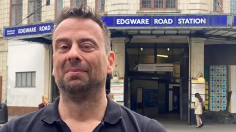 Head and shoulders image of Ben Thwaites, a man with dark brown hair and a beard. He looking into the camera. Behind him is Edgware Road Station in London. The station has a blue sign above it.
