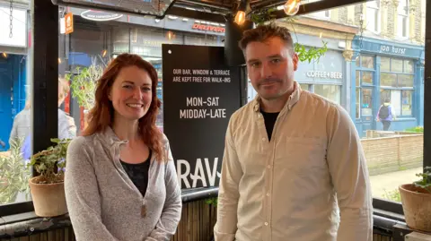 Imogen and Kieran Waite standing in their outdoor seating area. Behind them is a sign advertising the restaurant. Imogen has long ginger hair, and Kieran has a white shirt on and short cropped brown hair