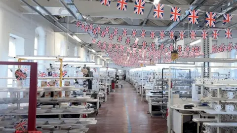 Inside a warehouse. The walls are painted white with big windows letting in the light. Strings of small Union Jack flags hang from the ceiling. A corridor goes down the middle of the room and on each side are rows of mugs and plates.