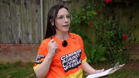 Richard Knights/BBC Naomi Woodford sostiene un portapapeles y un bolígrafo y habla a través de un micrófono que lleva adherido a su camiseta naranja. Su camiseta tiene "Campaña contra la vida miserable" a través de su frente. 