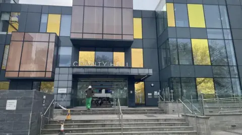 A modern-looking council building. There are steps leading to the entrance which has sliding doors. A person is leaving the building and walking towards the camera.