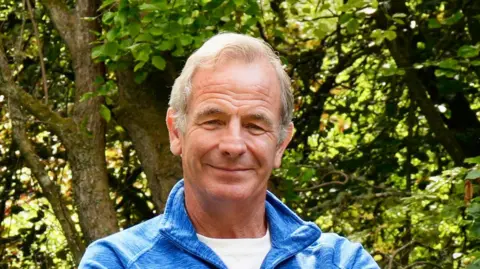 Robson Green. He is wearing a blue fleece and has grey hair. He is smiling at the camera.