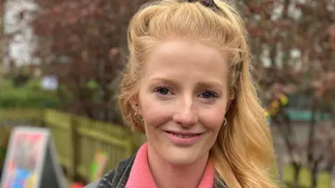 BBC Hannah Spencer has long blonde hair and is wearing a black denim jacket and pink sweater. She is standing outdoors in a playground.