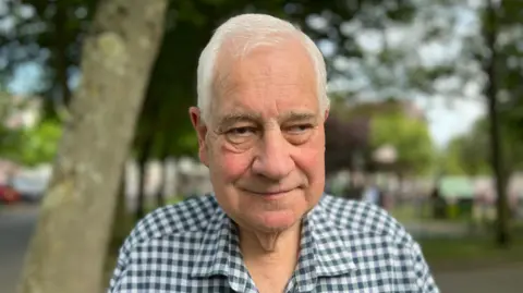 A man with white hair and a checked green and white shirt is looking into the camera. Behind him there are trees and some tarmac and grass.