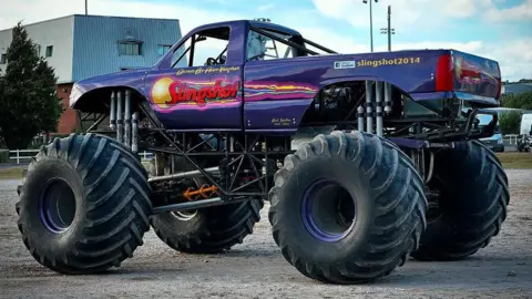 A purple monster truck is driving on a field. It has the word 'Slingshot' on the side of the truck.